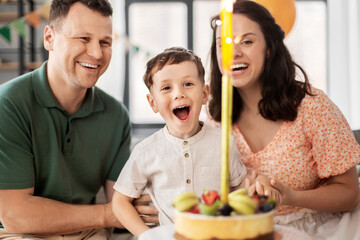 Wall Mural - family, holidays and people concept - portrait of happy mother, father and little son with firework candle burning on birthday cake sitting on sofa at home party