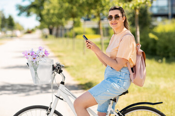 Poster - people, leisure and lifestyle - happy young woman with smartphone, backpack and flowers in basket of bicycle on city street