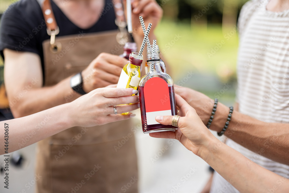 People clinking bottles with a strong alcohol drinks on a picnic outdoors, close-up. Holding bottles of liqueur or tinctures - obrazy, fototapety, plakaty 