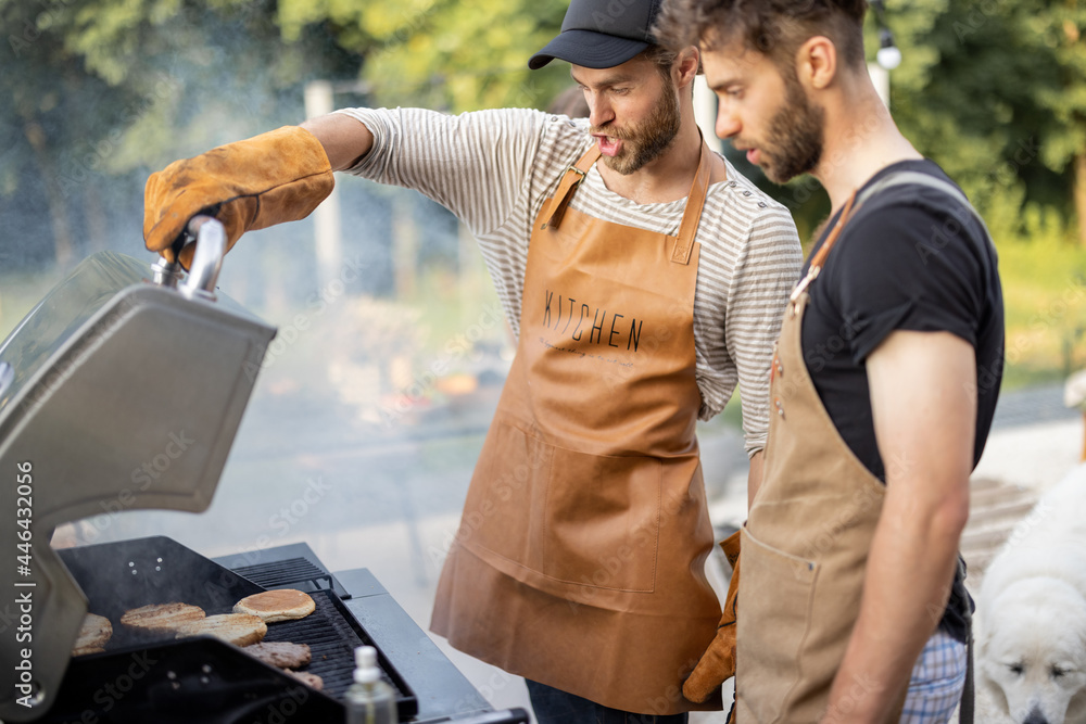 Two handsome male friends have fun while grilling meat and buns for burgers on gas grill at backyard on nature - obrazy, fototapety, plakaty 