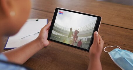 Wall Mural - Composite of woman sitting at table with face mask, watching rugby match on tablet