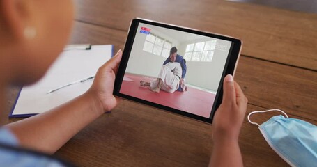 Wall Mural - Composite of woman sitting at table with face mask, watching judo match on tablet