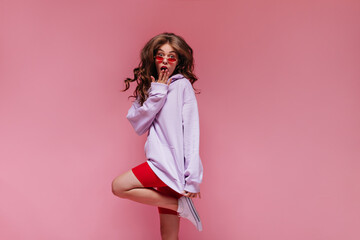 Wall Mural - Brunette curly woman in red sunglasses, purple hoodie and cycling shorts jumps on isolated. Cool girl looks surprised on pink background.