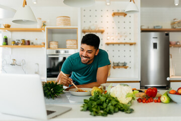 Happy African American man cooking healthy dinner at home. He is following a video tutorial on the laptop.