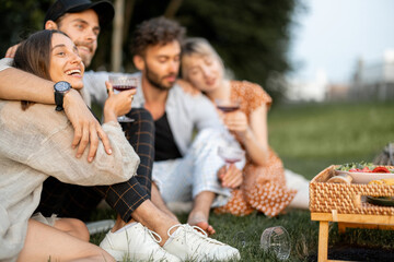 Young friends sit together and hug, talk and drink wine in a close and friendly atmosphere on a picnic in the evening