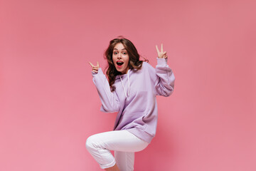 Wall Mural - Pretty brunette woman in oversized sporty style outfit looks surprised and shows peace sign. Cool curly girl in purple hoodie moves on pink isolated background.
