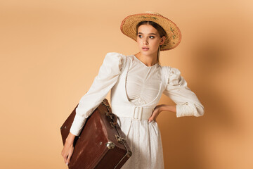 young woman in white dress and straw hat holding suitcase and posing with hand on hip on beige