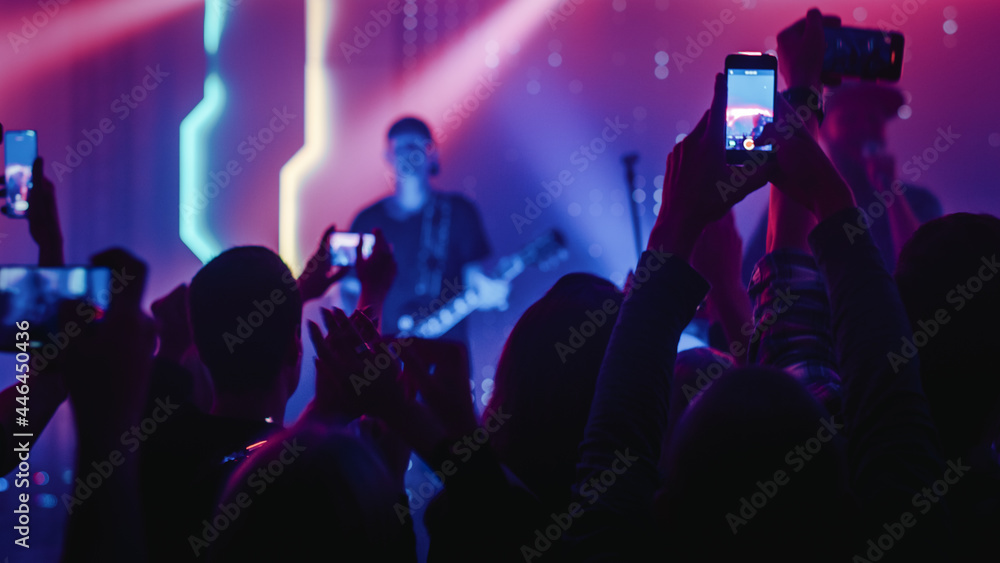 Rock Band with Guitarists and Drummer Performing at a Concert in a Night Club. Front Row Crowd is Record Video on Their Mobile Phones. Party in Front of Bright Colorful Strobing Lights on Stage.  - obrazy, fototapety, plakaty 