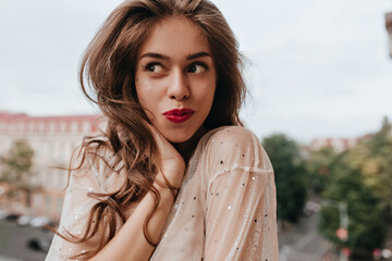 Wall Mural - Tanned brunette woman in white shirt poses in good mood. Pretty cheerful girl with red lips poses outside with smile.