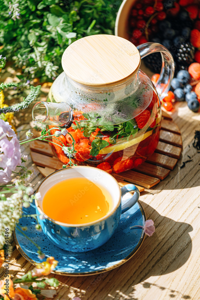 natural aromatic fruit tea in a transparent teapot and in a cup among berries and flowers. warming aromatic tea with a deep aroma of berries and wildflowers. - obrazy, fototapety, plakaty 