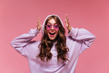 Wall Mural - Brunette woman in purple oversized hoodie looks into camera on pink background. Young teen girl in sunglasses poses on isolated.