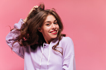 Wall Mural - Cool young curly woman in purple hoodie smiles widely. Brunette girl in sporty top touches hair and poses on pink isolated background.