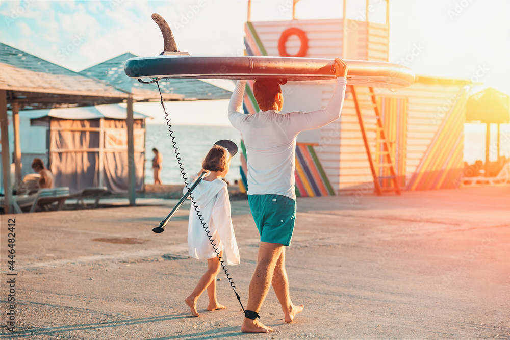 Father and daughter are walking along the beach. Man holding a sup board. Back view. Sunset light. Summer family vacation - obrazy, fototapety, plakaty 