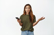 Frustrated middle aged woman has problem with mobile phone, looking complicated and confused, shrugging puzzled, holding smartphone, standing against white background