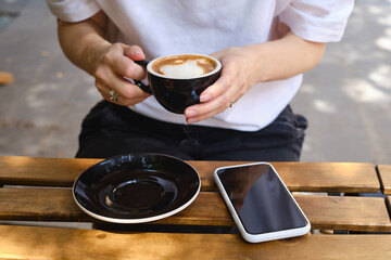 Girl drinks coffee from a black cup and uses a smartphone. A woman is holding a mobile phone, sending a text message or using an application on her mobile phone. Drink cappuccino or latte in a cafe