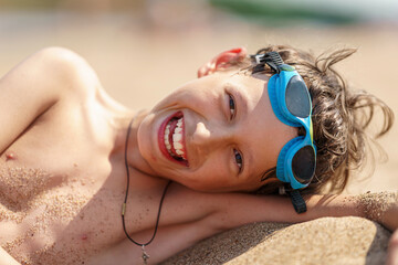 Wall Mural - Portrait of a happy boy in swimming glasses lying on the bank of a river in summer on a sunny day. A child enjoys a summer children's holiday on the lake shore. Active recreation.