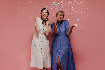 Wall Mural - Fashionable two women with short hairstyle in midi cool dresses smiling and posing with confetti on isolated background..