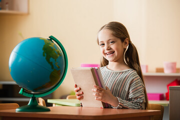 Wall Mural - pleased girl holding books while sitting near globe in montessori school