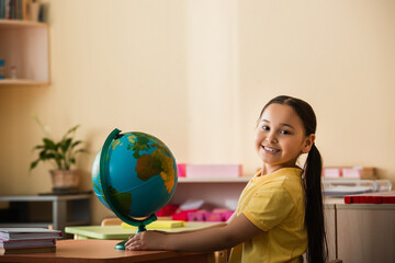 joyful asian girl smiling at camera near globe in montessori school