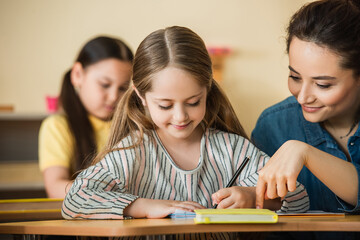 Wall Mural - teacher pointing near smiling kid writing in notebook and asian girl on blurred background