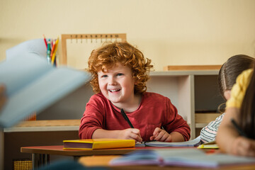 cheerful boy writing in notebook near blurred teacher and girls in classroom