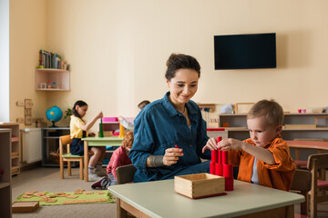 Wall Mural - young teacher smiling near boy making tower from wooden cylinders near kids on blurred background