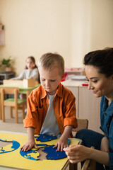 Wall Mural - boy combining earth map puzzle near smiling teacher and girl on blurred background