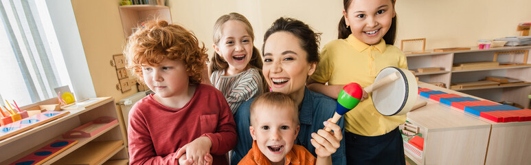 Wall Mural - cheerful teacher looking at camera while playing music with interracial kids, banner