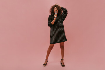 Poster - Wonderful woman with short curly hairstyle in modern striped dress with long sleeve and black heels singing and holding microphone on pink backdrop..