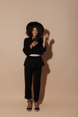 Wall Mural - Full length photo of wavy haired lady in hat, pants, black heels and shiny dark sweater with light belt looking into phone on beige backdrop..