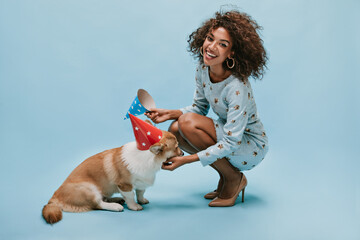Wall Mural - Beautiful lady with brunette hairstyle in blue clothes and heels posing with corgi in holiday cap on isolated background..