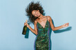 © Look! - Joyful woman with beautiful makeup in green charming dress playing with her curly hair and holding bottle with champagne on blue backdrop..
