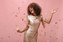 Happy Woman With Wavy Dark Hair Style In Shiny Trendy Clothes Rejoicing, Holding Glass With Champagne And Posing With Confetti On Pink Backdrop..