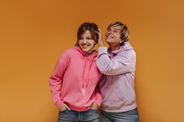 Cheerful two women with short hair in wide stylish hoodies and cool jeans smiling and having fun on orange isolated background..