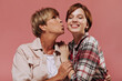 © Look! - Stylish woman with short blonde hairstyle in beige clothes kissing on cheek smiling girl in plaid outfit on pink background..