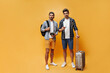 © Look! - Full-length portrait of brunet handsome men in colorful shorts, checkered shirts and white t-shirts hold suitcase, tickets and retro camera. Travelers pose on orange background.