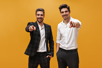 Good-humored young brunet businessmen in white shirts and black pants point at camera abs smile on isolated orange background.