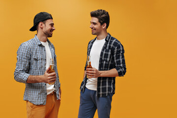 Wall Mural - Charming brunet bearded men in checkered stylish shirts and white t-shirts talk, hold beer bottles and smile on orange background.