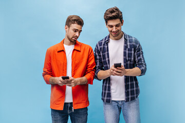 Wall Mural - Handsome young men in checkered shirt and orange jacket hold phones and pose on isolated blue background.