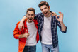 © Look! - Cool young bearded man in orange jacket and brunet guy in checkered shirt smile, show thumb up and ok sign on blue background.