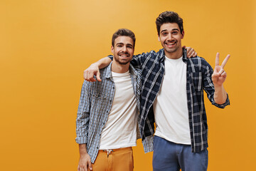 Cheerful young men in plaid blue shirts, white t-shirts and colorful pants pose on orange background in great mood and smile.