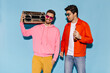 © Look! - Cheerful guy in yellow shorts and pink hoodie smiles sincerely and holds record player. Man in orange jacket and jeans holding beer bottle on blue background.