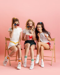 Three young women in retro 90s fashion style, outfits watching movie isolated over pink studio background. Concept of eras comparison, beauty, fashion and youth.