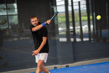 Man playing padel in a blue grass padel court indoor - Young sporty boy padel player hitting ball with a racket