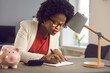 © Studio Romantic - Positive millennial african american business woman carefully writes notes sitting near calculator and piggy bank. Woman checks the bills, keeps track of expenses and plans the budget.