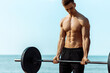 Muscular man training on the beach by the sea, doing exercises with a barbell for biceps, strong male press with a naked torso, sports concept