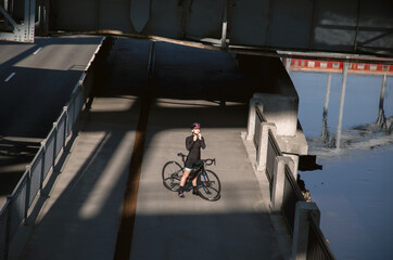 Wall Mural - Young woman athlete illuminated by the rays of the sun poses on the embankment after a morning workout on a bike. Go in for sports in the city. Sports paraphernalia.