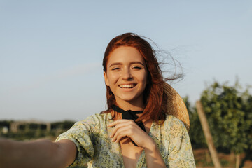 Wall Mural - Young woman with freckles, red hair and black bandage on neck in printed green clothes smiling and taking photo outdoor..