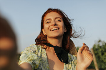 Wall Mural - Cheerful woman with red hairstyle and black bandage on neck in summer yellow and green dress laughing and making selfie outdoor..