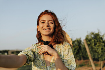 Wall Mural - Cheerful young girl with foxy hairstyle, straw hat and dark bandage on neck smiling and looking into camera outdoor